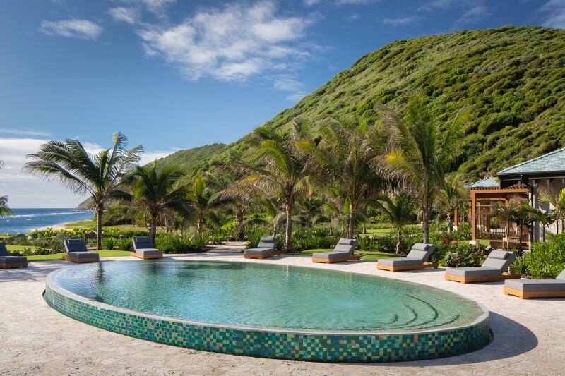 Shallow oval pool at Peter Island Resort, with several short palm trees, empty brown lounge chairs, and green hill at right