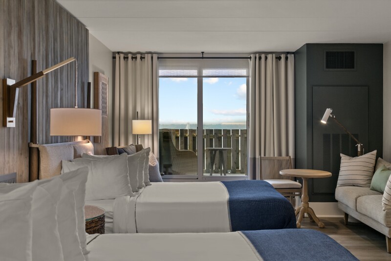 A guest room with two gray beds, wood paneling, and views of water outside glass door leading to small balcony