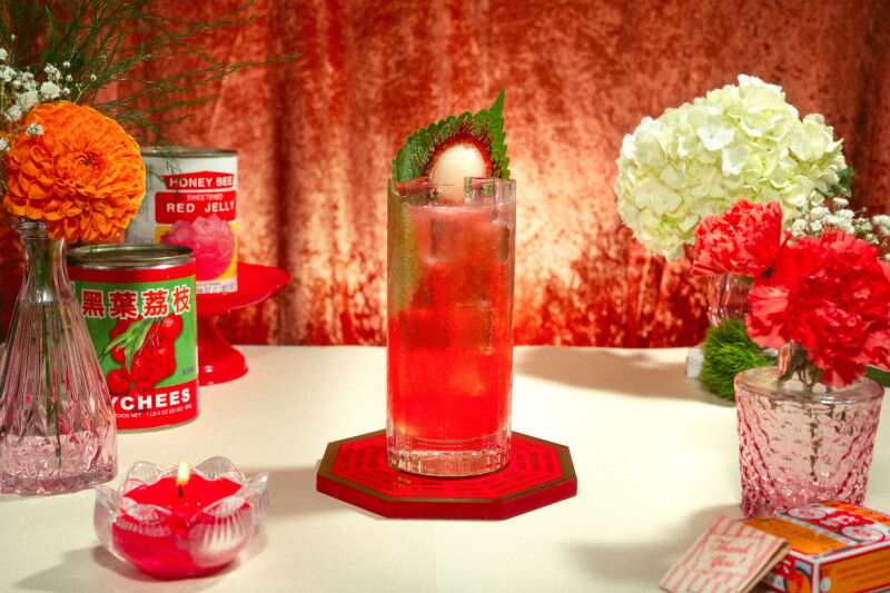 Tall red cocktail garnished with lychee and a fresh herb leaf, surrounded by canned lychees, orange and white flowers, and a small red candle.