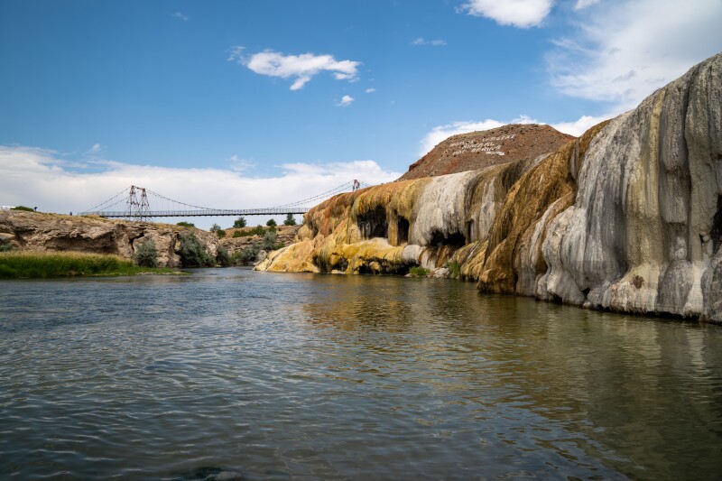 A suspension bridge in distance next to travertine stone cliffs beside expanse of water