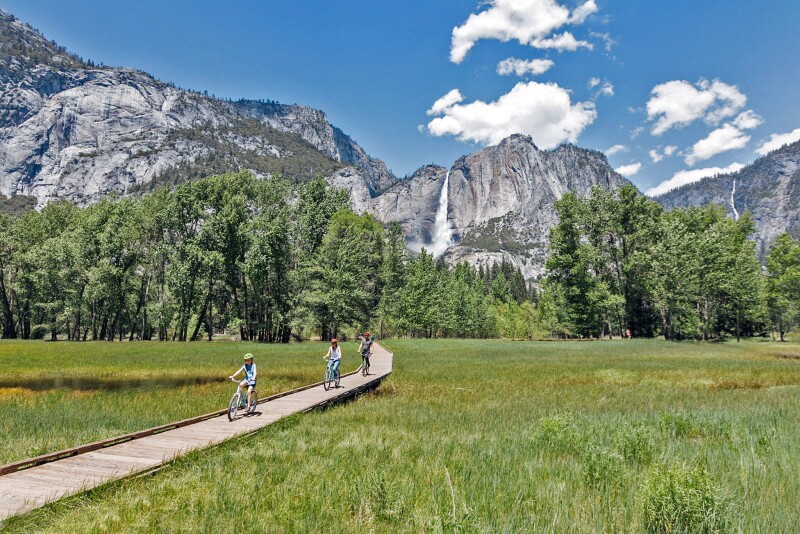 Two adults and a child bike on a wooden path that cuts through the green grass in the mountain valley. Behind them, green trees and large gray mountains loom.