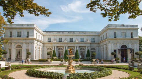 The exterior of an ornate white mansion with manicured gardens and a fountain.