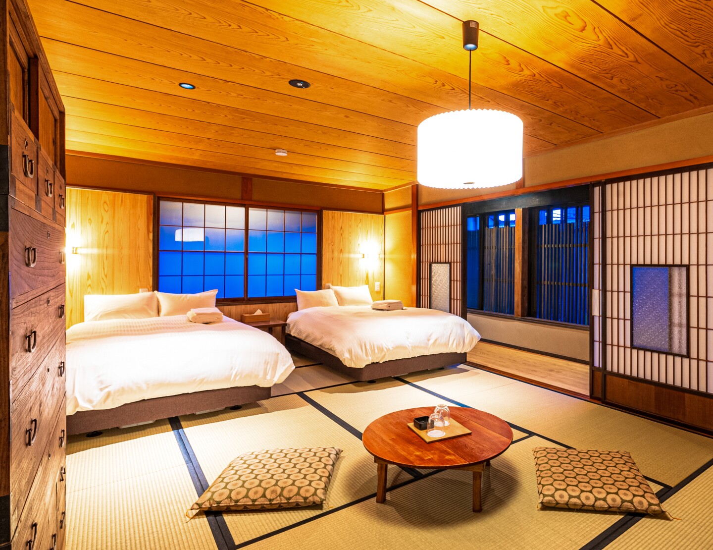 A guest room at Nipponia Sado, with tatami floors, two white beds, wooden ceiling, and windows