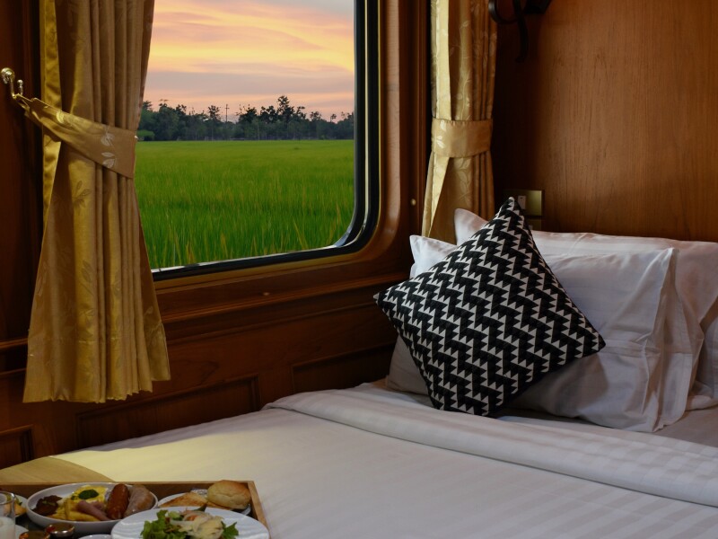 A Premium Suite on the Blue Jasmine Train Journey with a double bed up against a window with gold curtains and a view of the Thai countryside