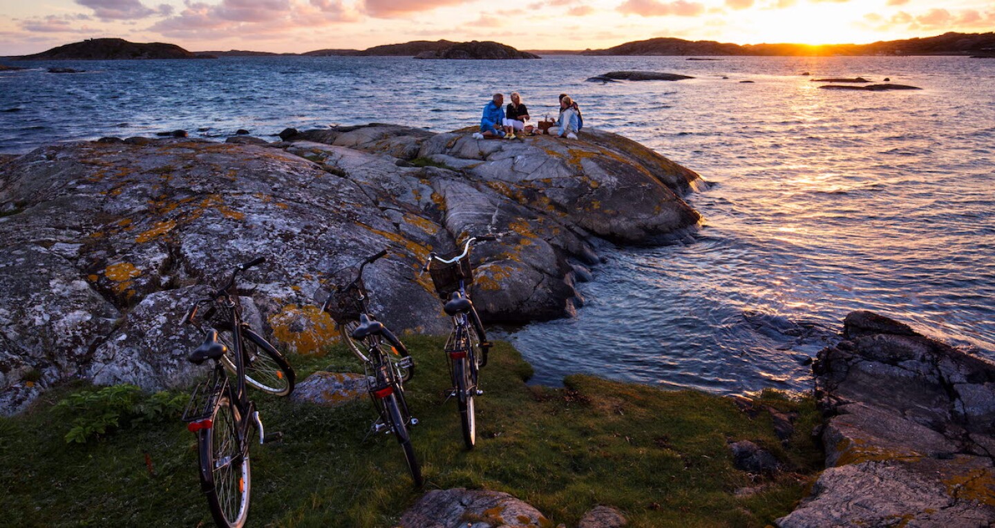 Four people eat at a picnic on a rock by the water. at sunset Their bicycles are waiting on the grass.
