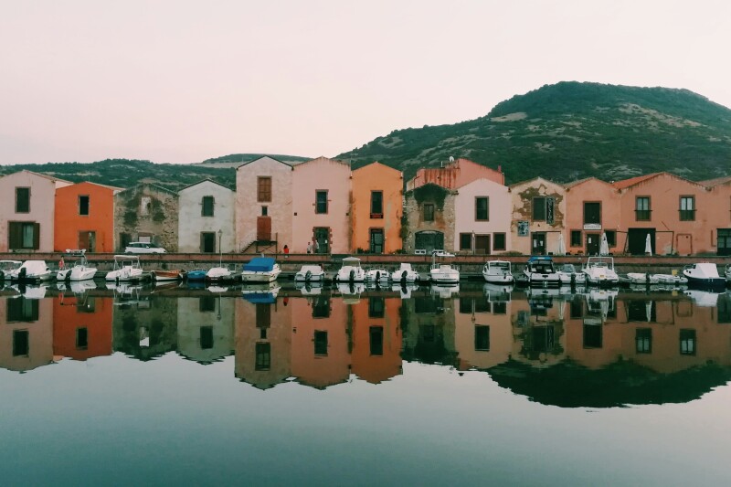 A row of two-story pink and pastel-colored buildings along waterfront lined with small boats, reflected in flat water