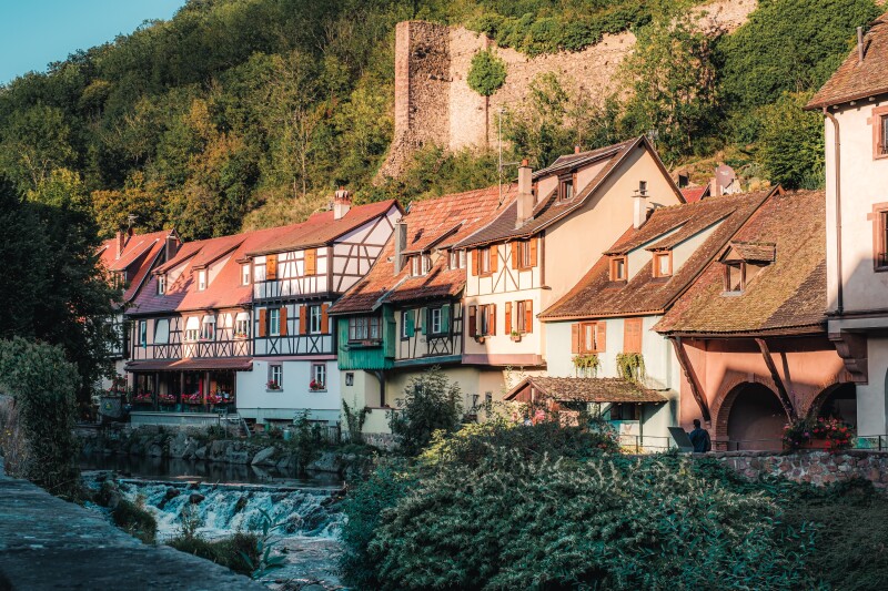 Row of traditional half-timbered houses beside stream, with old wall and hill in background