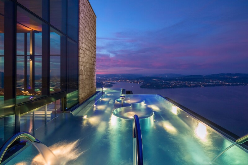 An infinity pool after sunset with built-in seating and jets shooting water like faucets, with a view out over an enormous lake