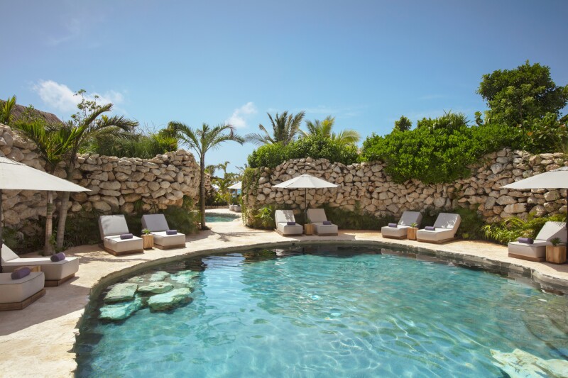 A freshwater cenotes at Impression Moxché by Secrets offer a welcome reprieve from the sun. Lounge chairs and umbrellas surround the cenote, which is bordered by a rock wall and foliage.