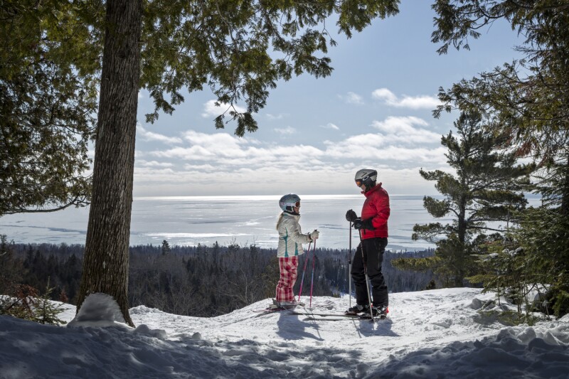 An adult and child wearing skis on the summit of Lutsen Mountains in Minnesota with Lake Superior in the background.