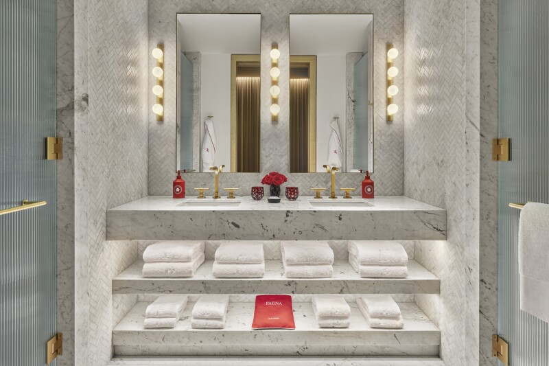 A bathroom in a guest room at Faena New York with a white and gray marble countertop and tiling and deep-red florals and soap dispensers