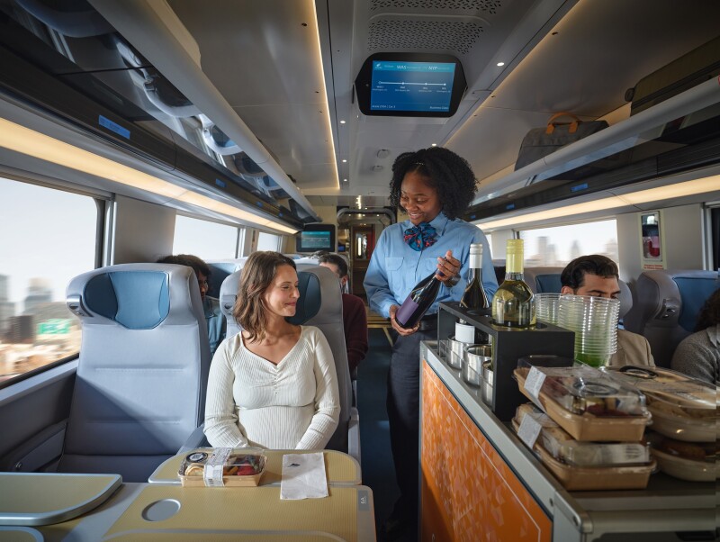 A woman seated on an Amtrak NextGen Acela train with a tray table topped with a to-go pack of food is being offered some wine from an attendant who is working the food and beverage cart service, and other passengers sit in the background