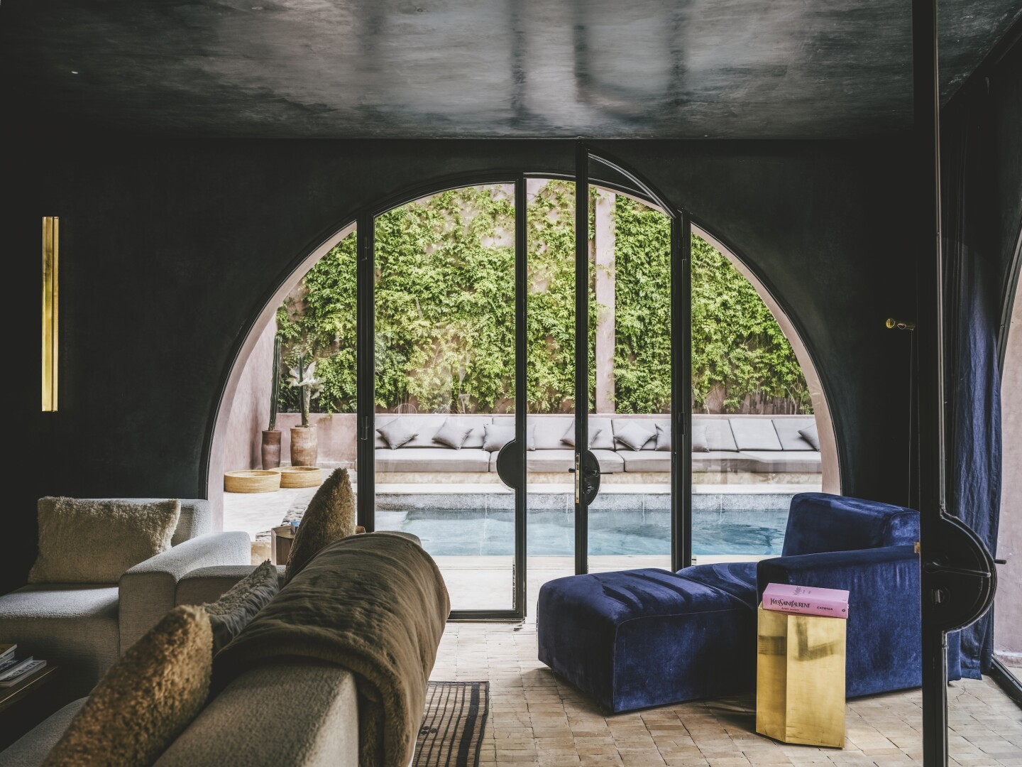 A modernist room with a green couch and a blue armchair leading outside thru an arched door to an outside swimming pool.