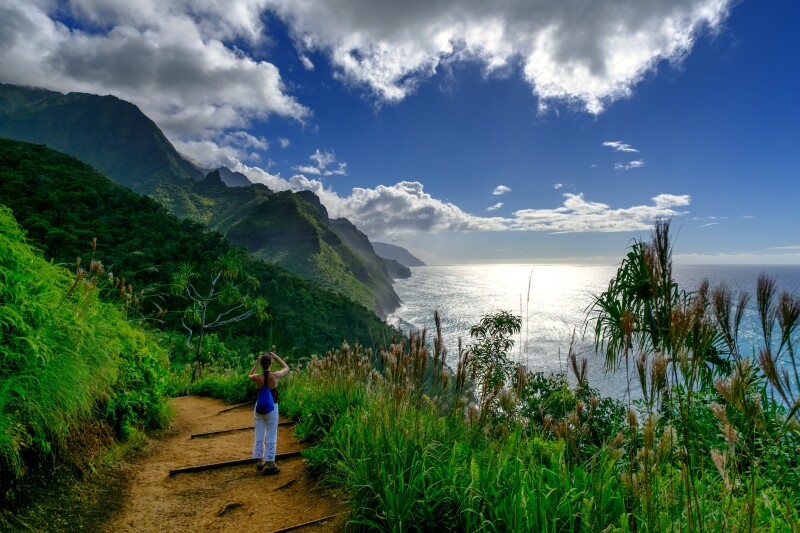A hiker taking a photo on a dirt path surrounded by tropical greenery, green mountains, and the ocean.