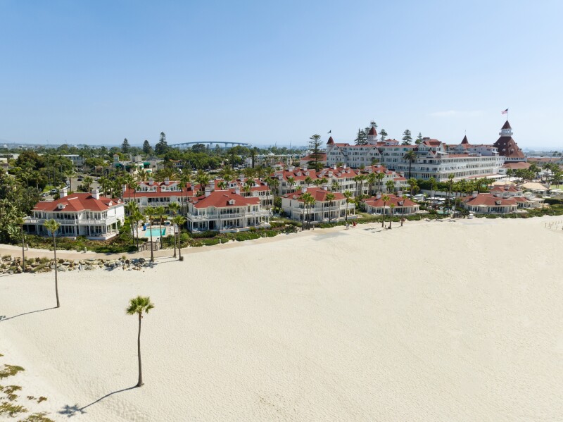 A wide view of the beach and grounds that comprise Hotel del Coronado, with white structures and dusty rose colored rooftops surrounded by palm trees.