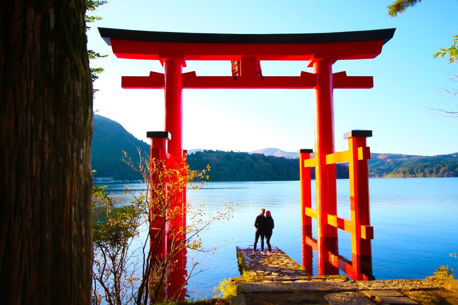 Two people standing under a traditional Japanese gateway by a lake in rural Japan.