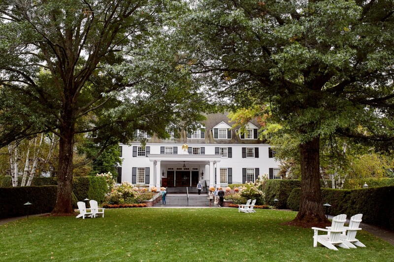 A white inn with dormer windows behind a green lawn with pairs of white Adirondack chairs