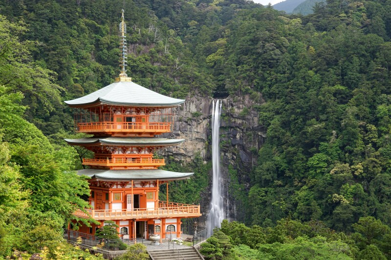 A four story orange-colored Japanese pagoda, with green forest and long waterfall in background