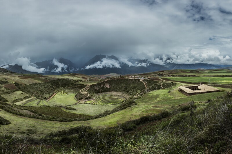 Aerial view of square building (at right) next to circular Inca ruins covered in grass, with cloudy, snowy Andes in distance