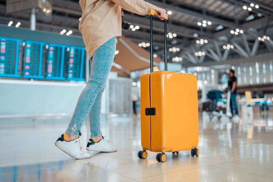 Bottom half of a woman wearing white sneakers, light-colored jeans, and a tan top, pushing a yellow hard suitcase through an airport
