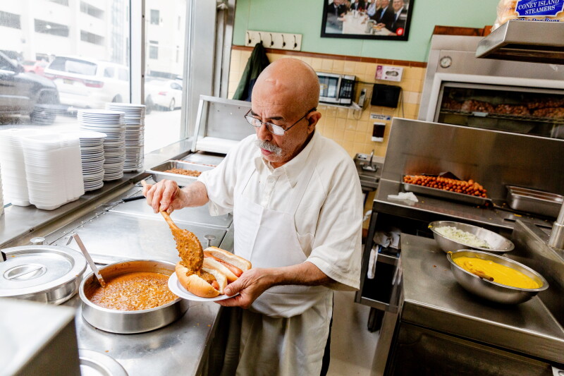 A chef in a white apron with glasses and a gray mustache pours chili from a large metal pot over two hot dogs at a Coney Island, or diner, in Detroit