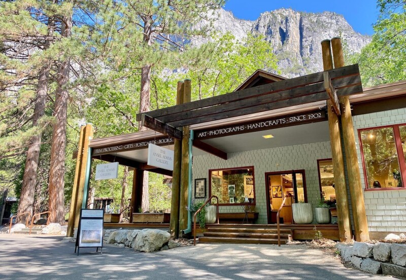 A small standalone building houses the Ansel Adams Gallery. Two wood pillars flank the front stairs. The awning states: Film Processing, Gifts, Postcards, Books, Art, Photographs, Handcrafts, Since 1902. Rocks frame the front entry and large trees are seen in the background.