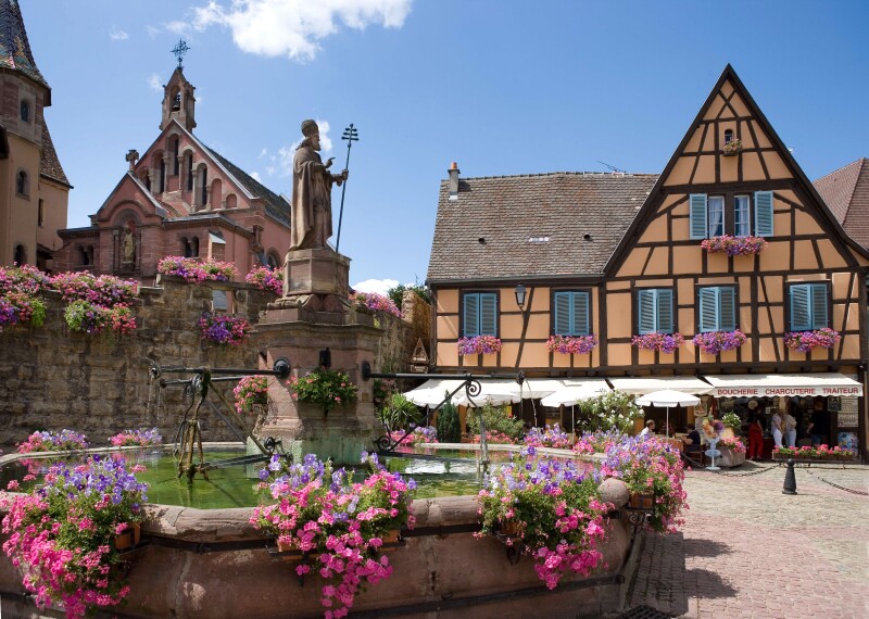 A public square in France, with pink flowers around fountain, a yellow half-timber building with blue shutters, statue of saint, and church.