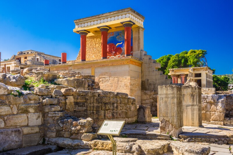 The Knossos complex in Crete, with a series of red columns sitting atop ruins and an image of a red bull painted on the exposed wall behind the columns