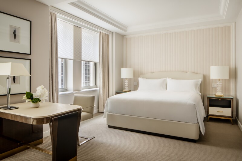 A guest room in white and cream, with desk, bed, nightstands, and two curtained windows