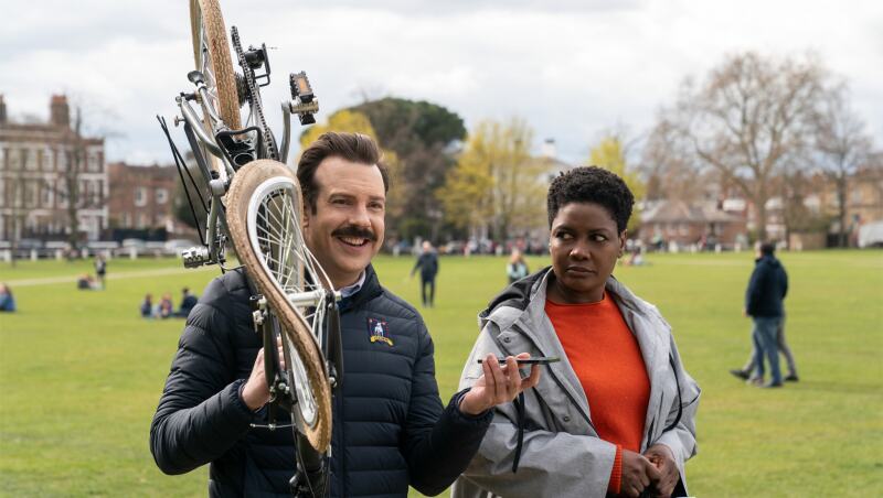 Ted Lasso carrying a mangled bike over his shoulder as he walks through a park with his doctor.