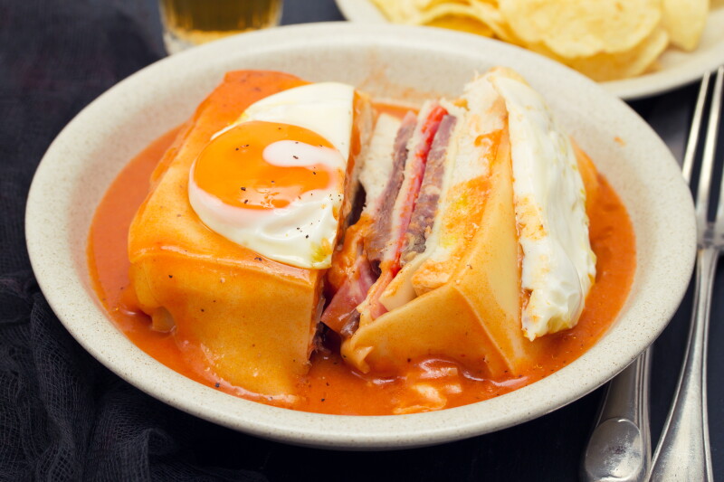 Un tazón blanco con un sándwich tradicional portugués en salsa de color naranja y huevo encima