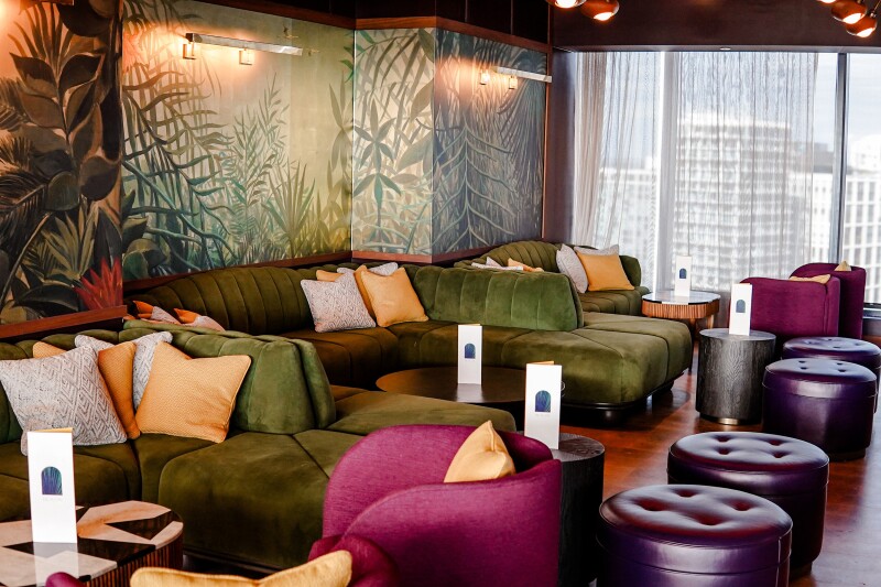 Bar with green and pink velvet couch seating and a palm-tree–inspired mural on the wall