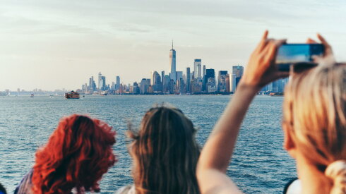 View of Manhattan skyline from the water. A woman with blond hair is holding up a camera to take a photo. Two other people are standing in front of her
