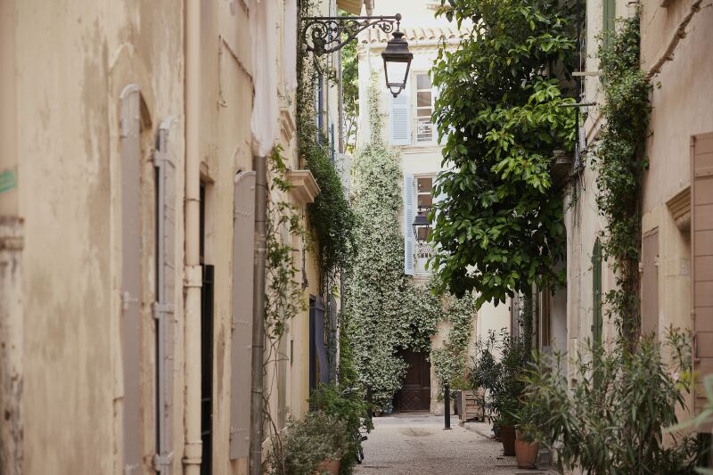 Sand-colored historic buildings with light-blue shutters and ivy on a street in Arles, France.