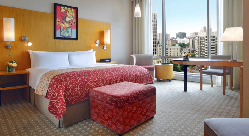 A bed with a red blanket and a red ottoman, a floral painting on the wall behind, and a desk by a large floor-to-ceiling window