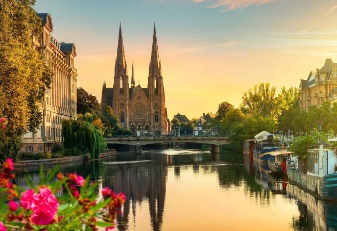 A view of a sunlit church on the Rhine river with pink flowers in the foreground in Strasbourg, France.