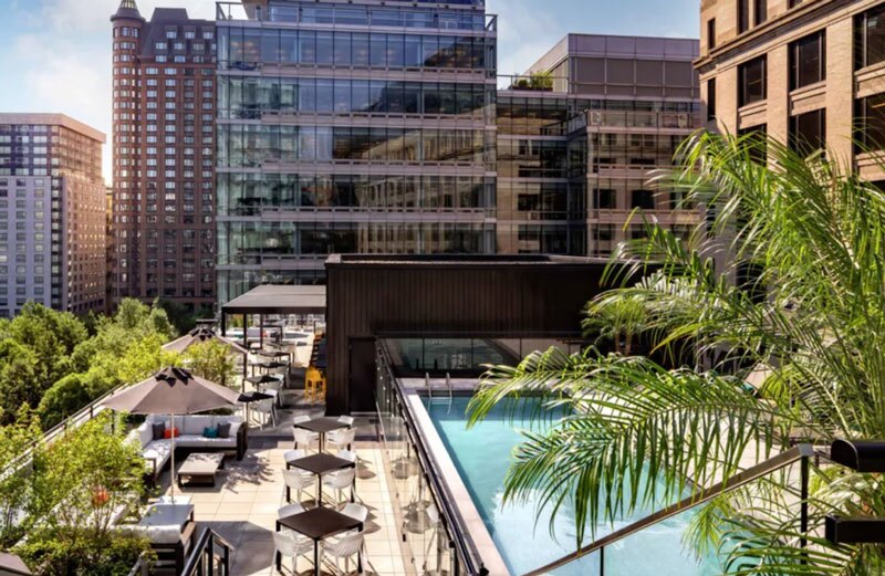 A rectangular rooftop pool surrounded by tables, umbrellas, and palm trees, with high-rises in the background