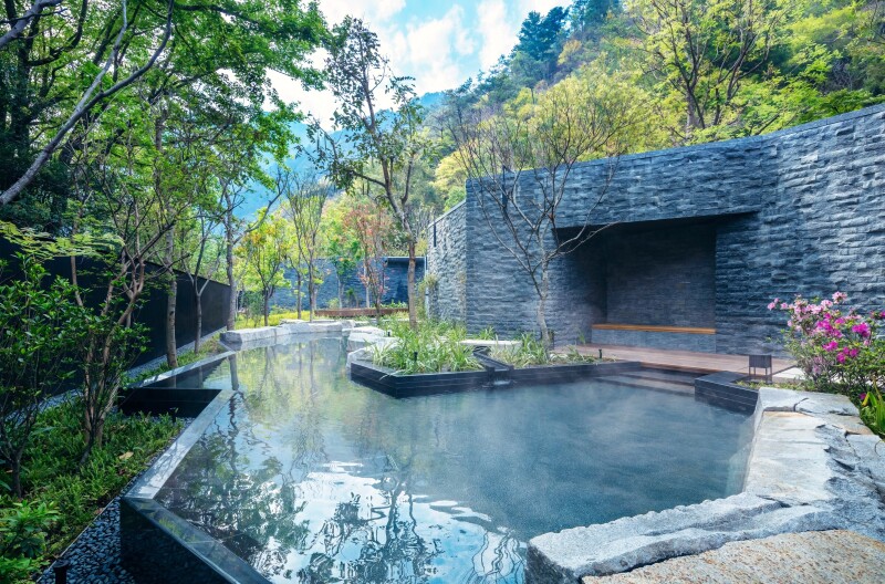 An outdoor hexagonal-shaped pool, with a grey stone wall behind it, surrounded by trees