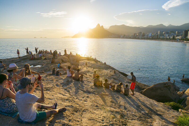Crowd of people gathers to watch the sunset on a rock. at Arpoador in what has become a popular daily tradition.