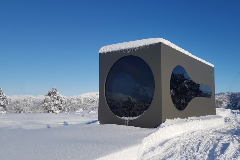 A rectangular, dark gray house with one large round and one oddly shaped window in a snowy field with trees in the distance