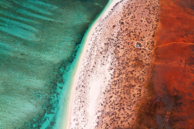 Aerial view of a beach with blue green waters on the left, a strip of white sand in the center, and red dirt to the right