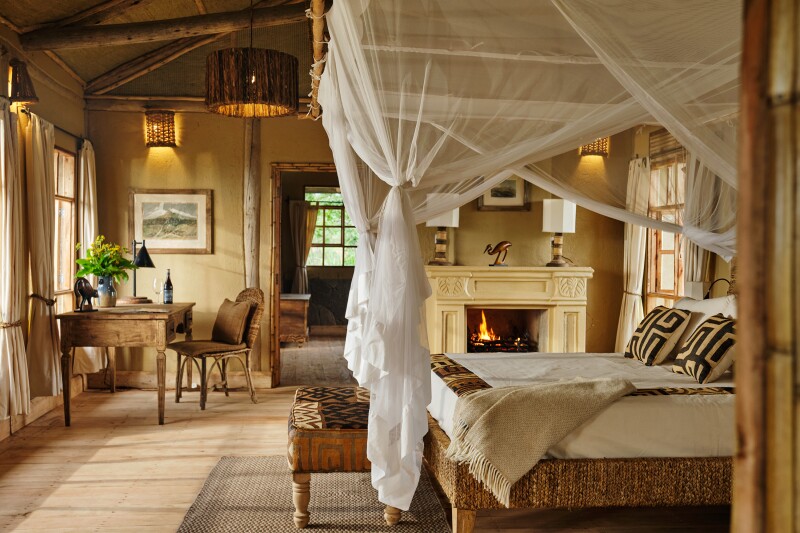 Interior of African safari lodge room, with four-poster bed surrounded by gauzy netting, brown and beige furnishings, patterned pillows, plus fireplace in background