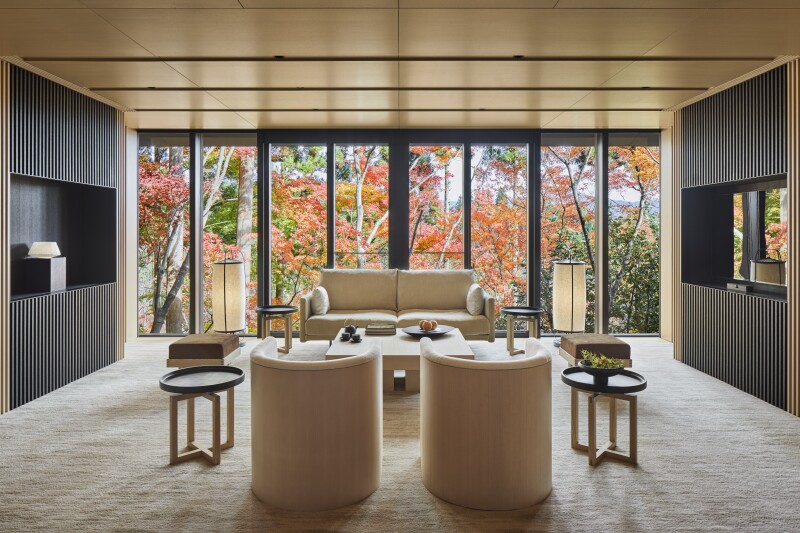 A beige room in suite at Aman Kyoto, with pair of curved chairs facing sofa and wall of windows overlooking fall foliage trees