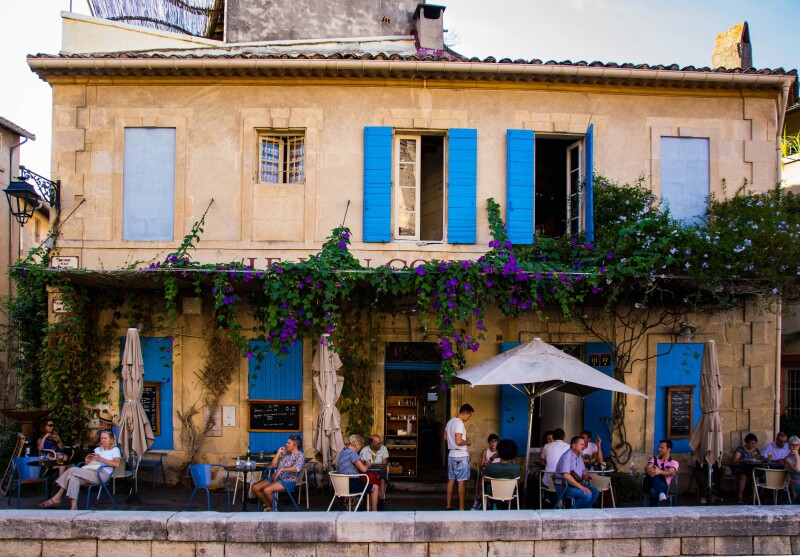 People sitting at an outdoor terrace of a restaurant or café in Arles, France, with blue shutters and a flowering vine lining the beige building