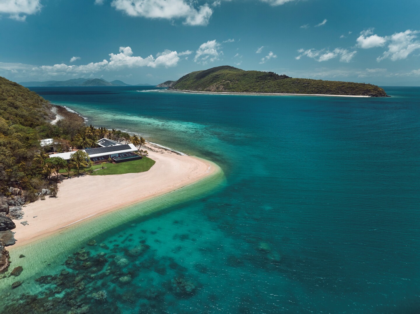 Aerial view of villa on a white-sand beach overlooking expansive clear waters with a hilly green island in the background