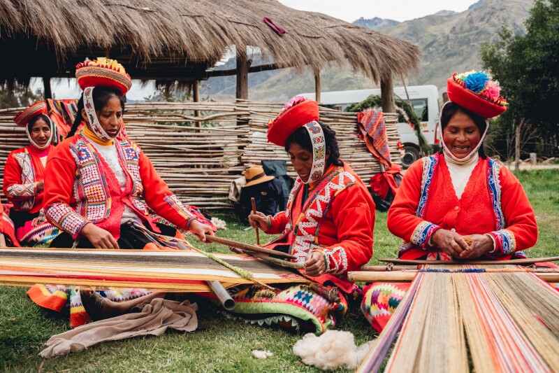 Several women from the Indigenous Huilloc Alto community demonstrate weaving outdoors in Peru; each wears a bowl-shaped red hat