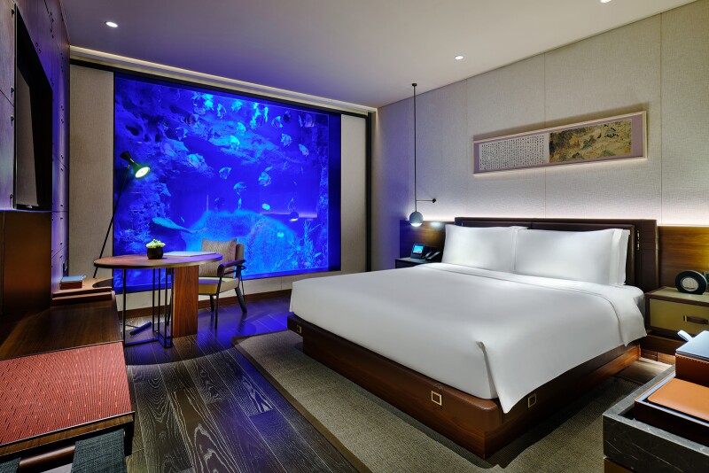 A bedroom with a table, lamp, and white bed, with large window looking into a fish-filled aquarium