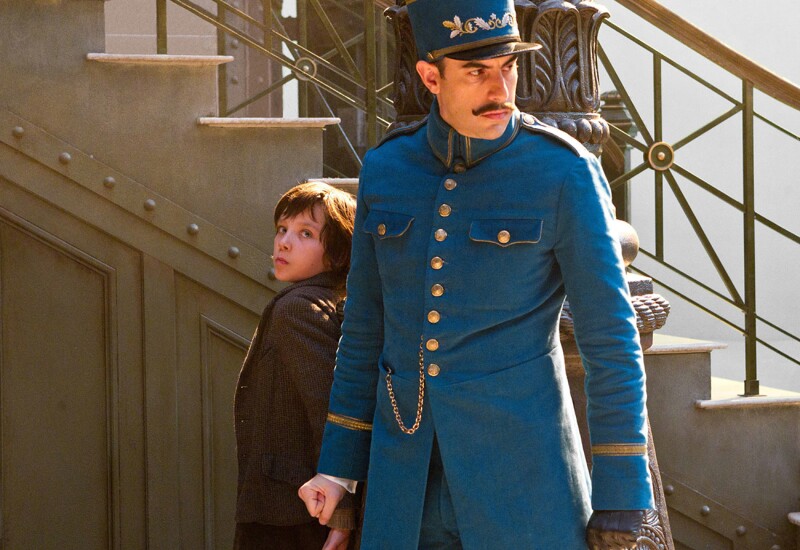 Asa Butterfield wearing a brown coat and Sacha Baron Cohen wearing a blue officer uniform stand in front of a staircase in the movie 'Hugo.'