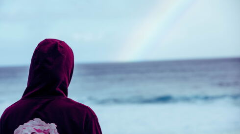 the back of a person in a burgundy hoody with a flower on it looking out across the ocean with a faint rainbow in the distance