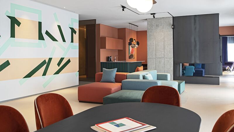 Lobby area. A conference table with brown, round chairs in the foreground. Brown-orange and light blue lounge chairs are in front of a reception desk. Rustic walls with abstract artwork.